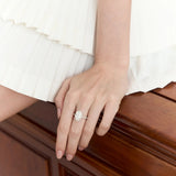 Hand wearing oval lab-grown diamond engagement ring on wood surface, white pleated skirt