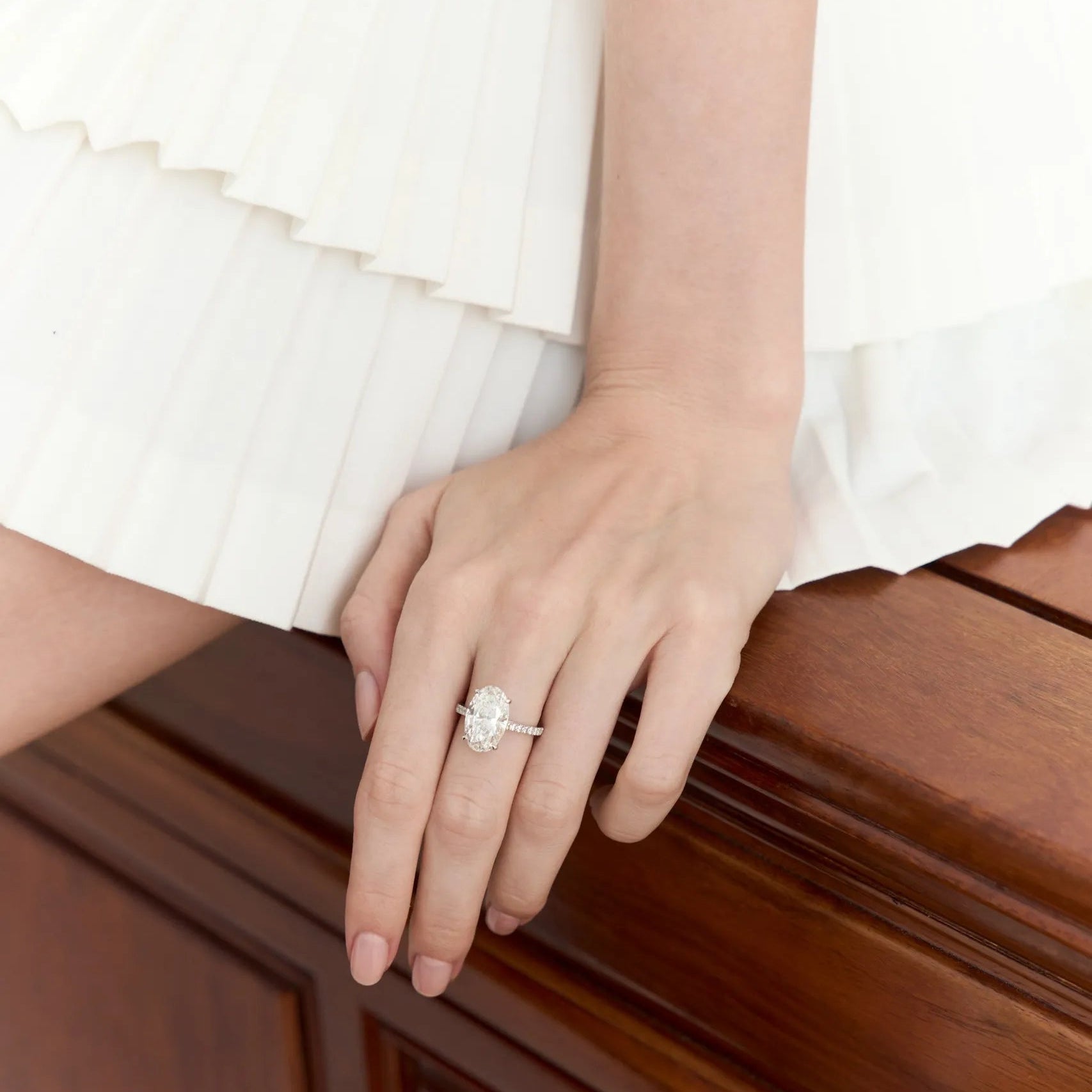 Hand wearing oval lab-grown diamond engagement ring on wood surface, white pleated skirt